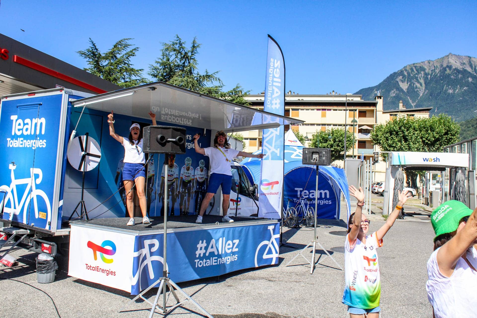 Camion podium Team TotalEnergies - deux animatrices sur la scène déployée face au public, montagnes en arrière-plan