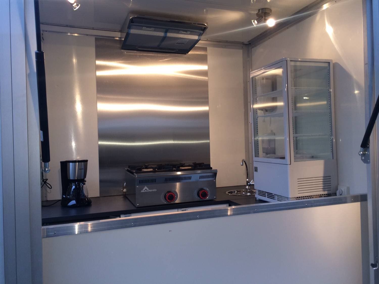 Intérieur de l'Aixam Mega aménagé avec plaque de cuisson Mainho, cafetière, vitrine réfrigérée et évier inox