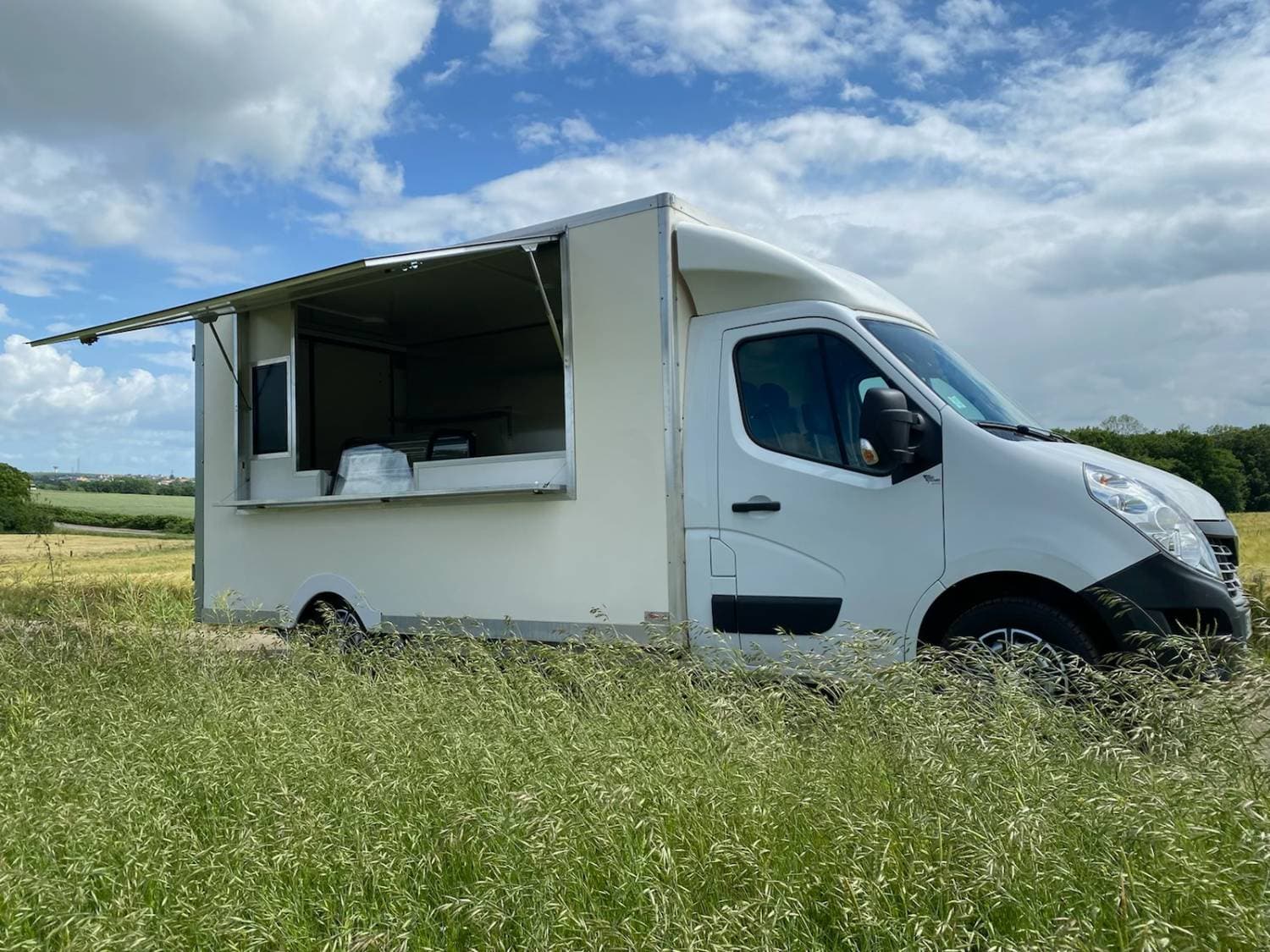 Camion traiteur blanc Renault Master volet ouvert côté service dans un champ de hautes herbes