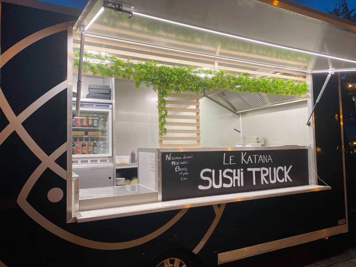 Food truck Le Katana Sushi Truck noir ouvert de nuit avec éclairage LED, végétation décorative et ardoise menu