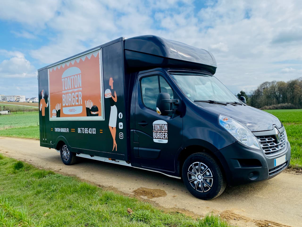 Food truck Tonton Burger noir et orange sur base Renault Master stationné dans un champ verdoyant