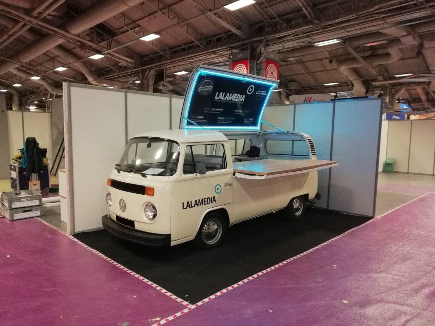 Combi VW blanc Lalamedia sur un stand de salon avec écran LED rétroéclairé et comptoir en bois - vue de face
